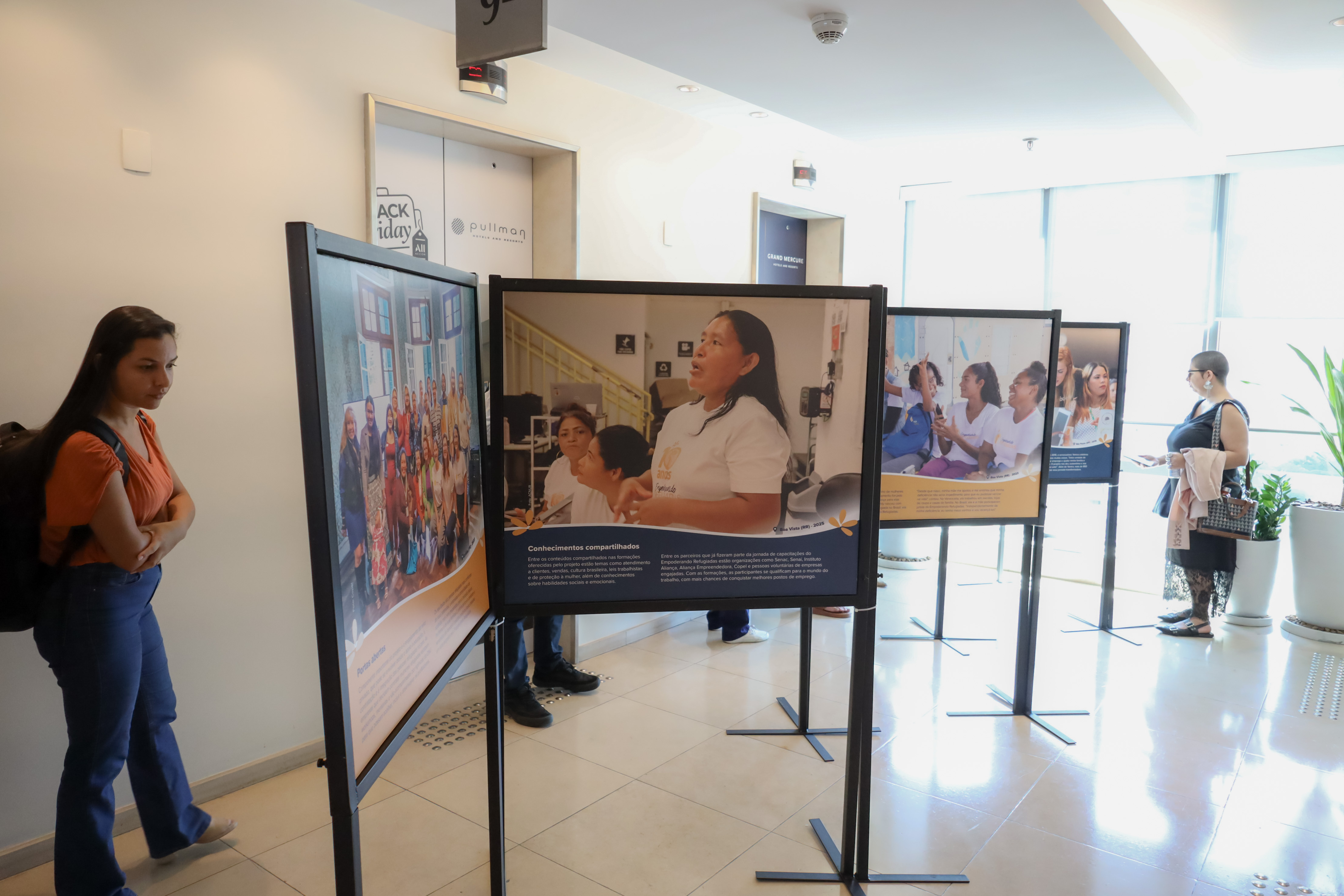 Visitantes observam uma exposi&ccedil;&atilde;o fotogr&aacute;fica instalada no hall de um pr&eacute;dio, com pain&eacute;is que mostram mulheres refugiadas em atividades de capacita&ccedil;&atilde;o e integra&ccedil;&atilde;o. As fotos est&atilde;o dispostas em suportes met&aacute;licos alinhados, enquanto pessoas passam e leem as legendas. O ambiente &eacute; claro e moderno, com piso claro e luz natural entrando pelas janelas.