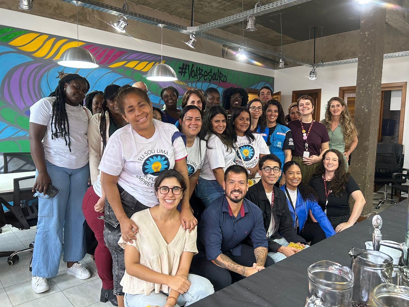 Grupo diverso de mulheres refugiadas e facilitadores reunidos em uma sala de forma&ccedil;&atilde;o, posando para uma foto coletiva. Elas sorriem e vestem camisetas do curso &lsquo;Eu Sou Uma Mulher Empreendedora&rsquo;. O ambiente &eacute; colorido, com mural art&iacute;stico ao fundo e mesas de trabalho, indicando um espa&ccedil;o de capacita&ccedil;&atilde;o e conviv&ecirc;ncia.