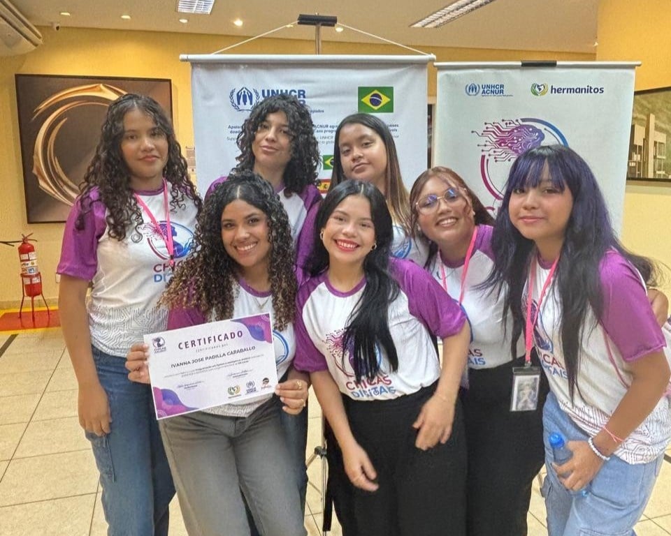 Grupo de pessoas usando camisetas do programa &lsquo;Chicas Digitais&rsquo; posando juntas em frente a dois banners com logos do ACNUR, Hermanoitos e a bandeira do Brasil. Uma das pessoas segura um certificado na m&atilde;o. O grupo est&aacute; em um ambiente interno bem iluminado, com paredes claras e obras de arte ao fundo.