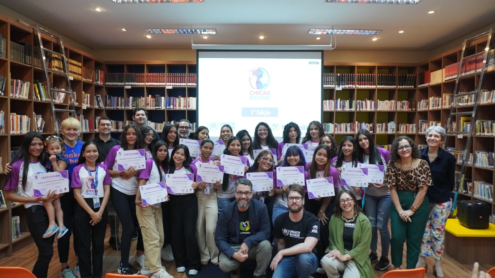 Grande grupo de pessoas reunidas em uma biblioteca para um evento do programa &lsquo;Chicas Digitais&rsquo;. A maioria est&aacute; posicionada em p&eacute;, lado a lado, segurando certificados com a marca do programa. Tr&ecirc;s pessoas est&atilde;o agachadas &agrave; frente do grupo. Ao fundo, h&aacute; uma tela de proje&ccedil;&atilde;o exibindo o logotipo do evento e estantes altas repletas de livros em toda a parede. O ambiente &eacute; iluminado e organizado, com cadeiras coloridas distribu&iacute;das nas laterais.