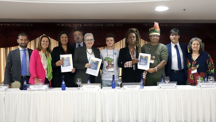 Grupo de pessoas posicionadas atr&aacute;s de uma mesa comprida coberta com toalha branca, em uma sala de eventos. Algumas pessoas seguram documentos impressos. H&aacute; garrafas de &aacute;gua e ta&ccedil;as sobre a mesa, al&eacute;m de placas de identifica&ccedil;&atilde;o com nomes. Ao fundo, cortinas em tons escuros e claros.