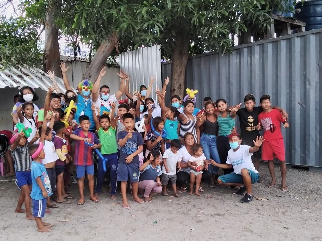 Grupo de crian&ccedil;as e adultos reunidos em espa&ccedil;o comunit&aacute;rio ao ar livre durante atividade recreativa, com bal&otilde;es e &aacute;rvores ao fundo.
