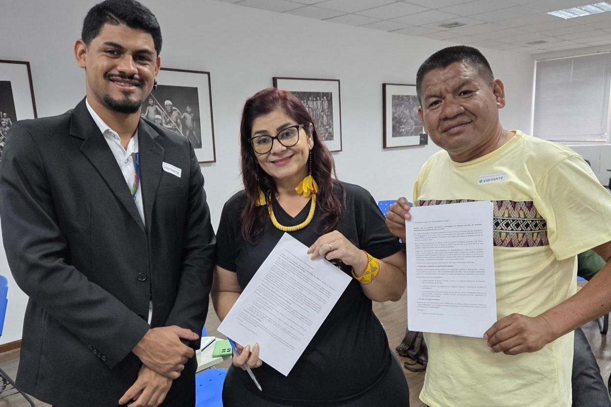 Tr&ecirc;s pessoas em ambiente interno posam lado a lado diante de uma parede com fotografias emolduradas. As tr&ecirc;s seguram documentos impressos nas m&atilde;os, voltados para a c&acirc;mera, em uma sala com cadeiras azuis e ilumina&ccedil;&atilde;o artificial.