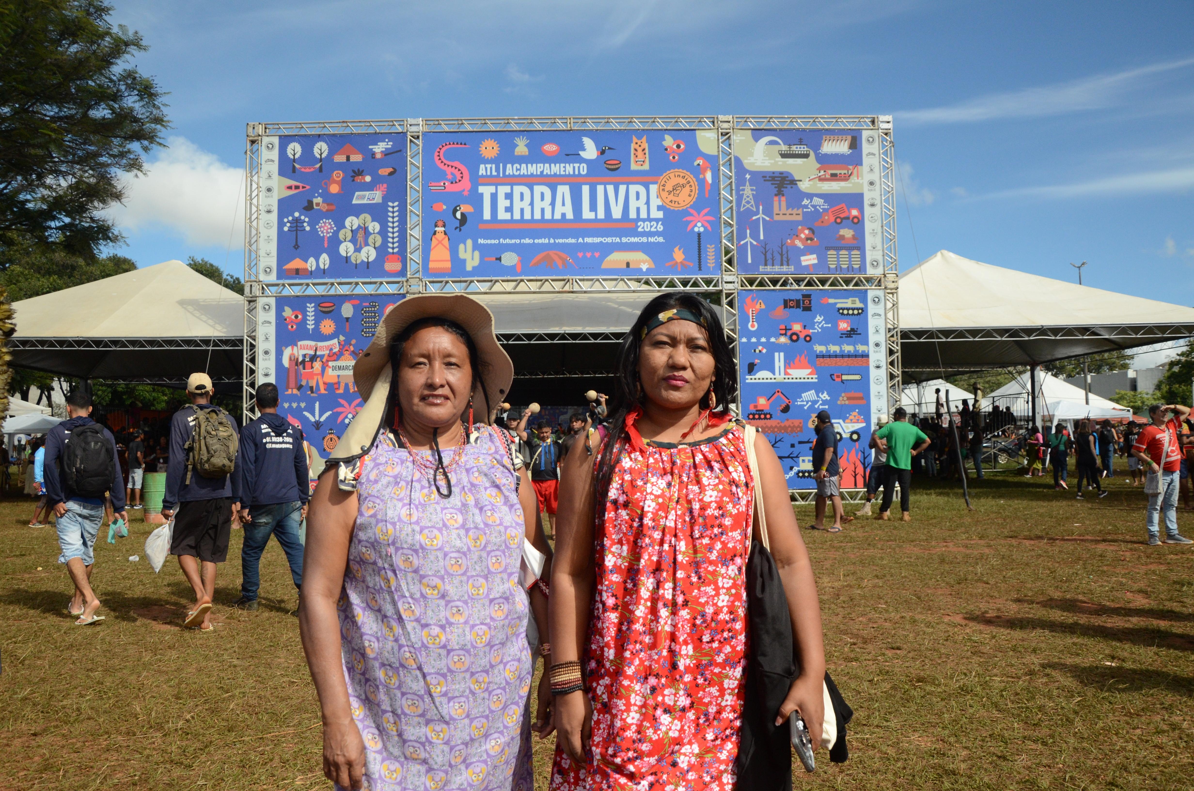 Duas mulheres em p&eacute; em &aacute;rea aberta de evento, em frente a uma entrada com estrutura met&aacute;lica e banner do &lsquo;Acampamento Terra Livre 2026&rsquo;. Ao fundo, tendas brancas, pessoas circulando e ilustra&ccedil;&otilde;es coloridas no painel principal.