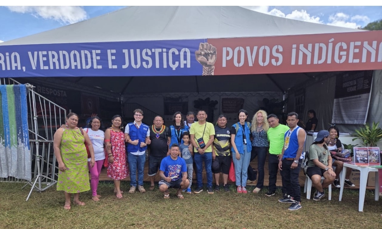 Grupo de pessoas reunidas em &aacute;rea externa, posando em frente a uma tenda com a faixa &lsquo;Mem&oacute;ria, Verdade e Justi&ccedil;a &ndash; Povos Ind&iacute;genas&rsquo;. As pessoas est&atilde;o alinhadas em p&eacute; e agachadas sobre um gramado, com mesas, materiais informativos e a estrutura da tenda vis&iacute;veis ao fundo.