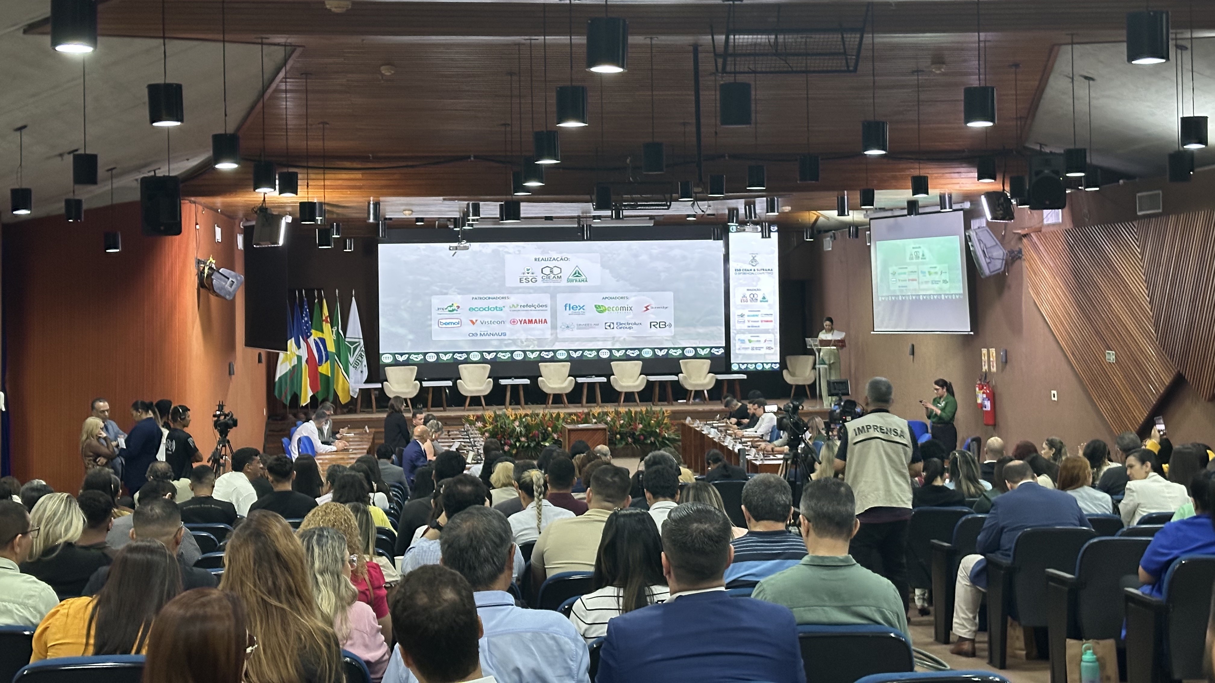 Audit&oacute;rio cheio com p&uacute;blico sentado assistindo apresenta&ccedil;&atilde;o em palco durante evento institucional, com tela de proje&ccedil;&atilde;o e bandeiras ao fundo.