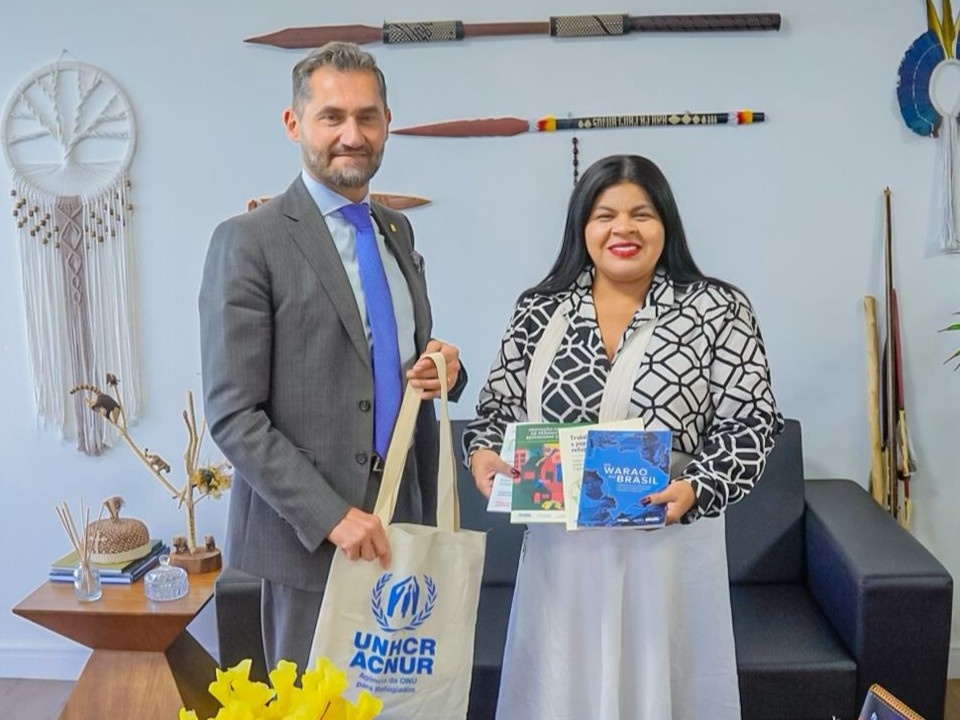 Homem de terno cinza e mulher ind&iacute;gena blusa estampada preta e branca posam sorrindo em um escrit&oacute;rio decorado com elementos ind&iacute;genas. O homem segura uma sacola do ACNUR, enquanto a mulher segura tr&ecirc;s livros coloridos. Atr&aacute;s deles, h&aacute; lan&ccedil;as, artesanatos de parede e objetos decorativos sobre uma mesa lateral.