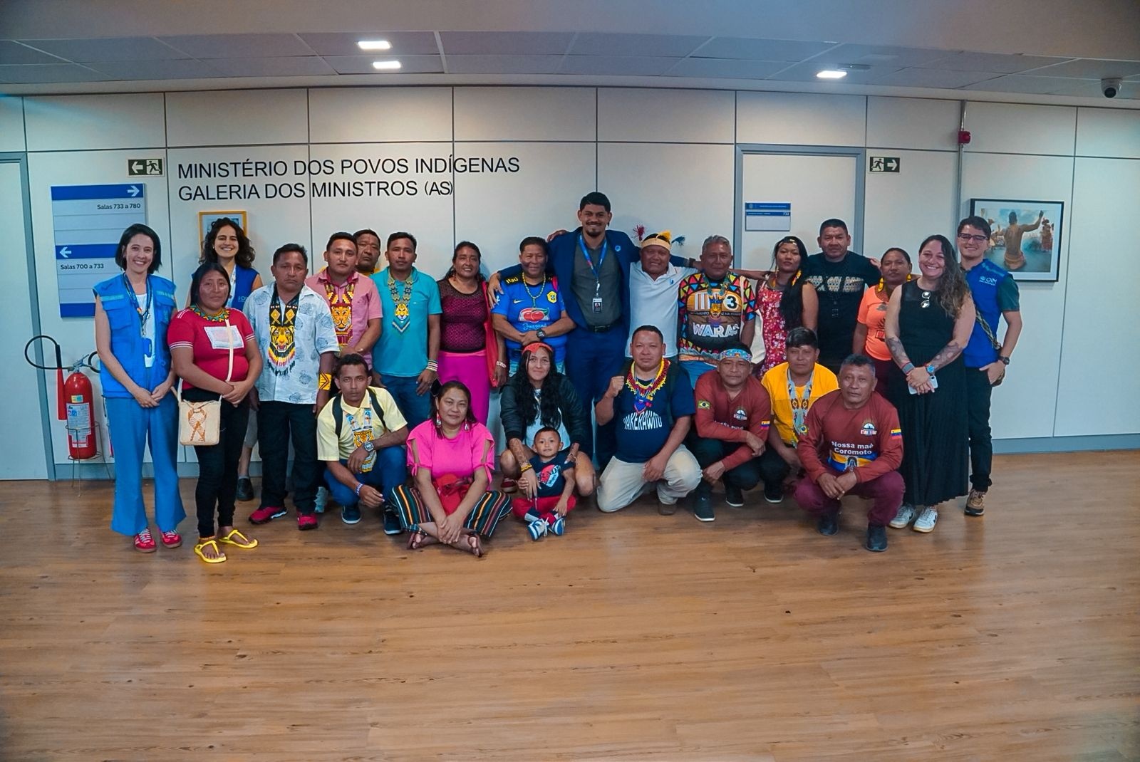 Grupo numeroso de pessoas posando para foto em ambiente interno, em frente a uma parede com a identifica&ccedil;&atilde;o &lsquo;Minist&eacute;rio dos Povos Ind&iacute;genas &ndash; Galeria dos Ministros (as)&rsquo;. As pessoas est&atilde;o dispostas em p&eacute; e agachadas, com um beb&ecirc; ao centro da primeira fila, em um espa&ccedil;o com piso de madeira e ilumina&ccedil;&atilde;o artificial.