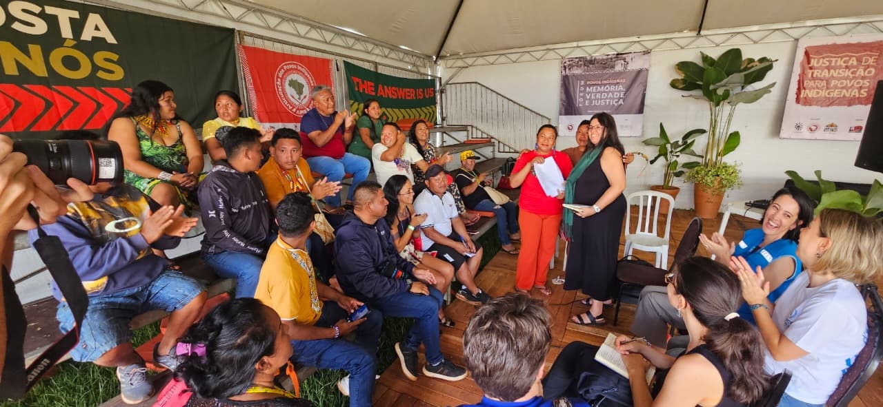 Grupo de pessoas reunidas em um espa&ccedil;o coberto com cadeiras dispostas em semic&iacute;rculo, durante uma atividade coletiva. Duas pessoas est&atilde;o em p&eacute; ao centro, uma delas segurando pap&eacute;is, enquanto as demais acompanham sentadas e aplaudem. Ao fundo, faixas com textos sobre justi&ccedil;a de transi&ccedil;&atilde;o, mem&oacute;ria e direitos dos povos ind&iacute;genas.