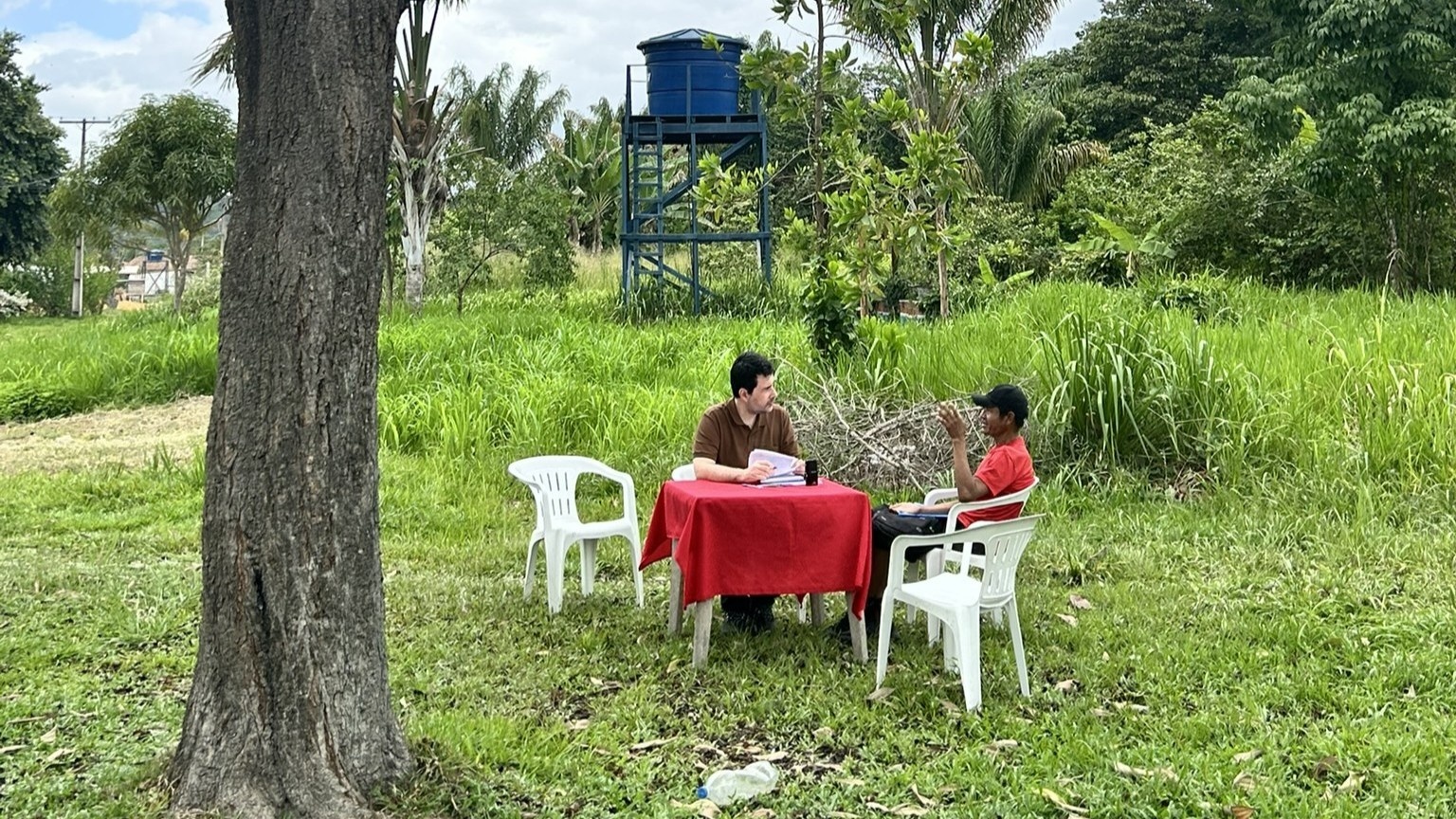 Duas pessoas sentadas em cadeiras pl&aacute;sticas conversam em uma mesa ao ar livre, em &aacute;rea verde, com vegeta&ccedil;&atilde;o densa e uma caixa-d&rsquo;&aacute;gua elevada ao fundo.