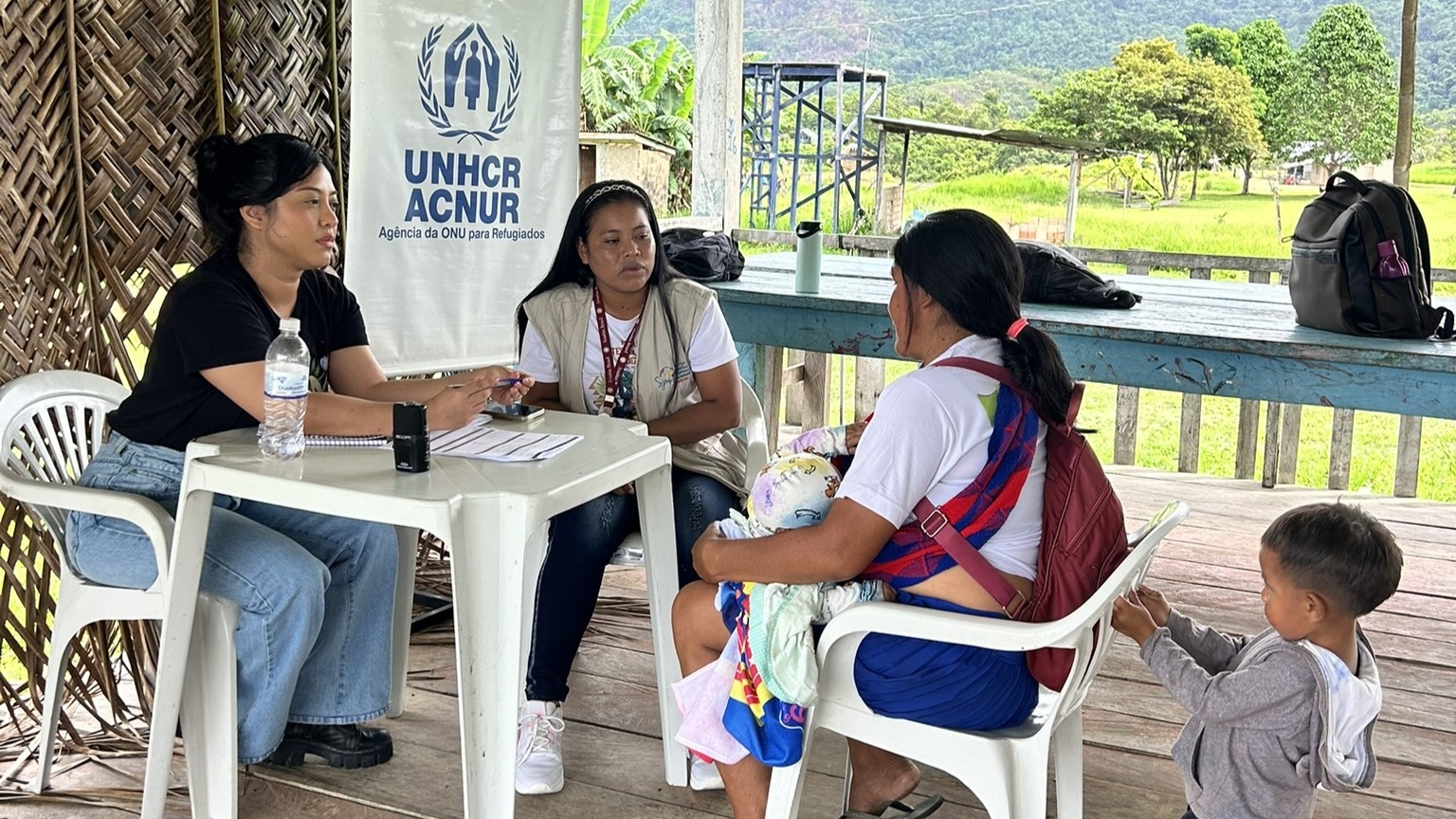 Atendimento individual realizado por integrantes do MJSP em mesa com formul&aacute;rios, em espa&ccedil;o comunit&aacute;rio, com banner institucional do UNHCR/ACNUR vis&iacute;vel ao fundo.
