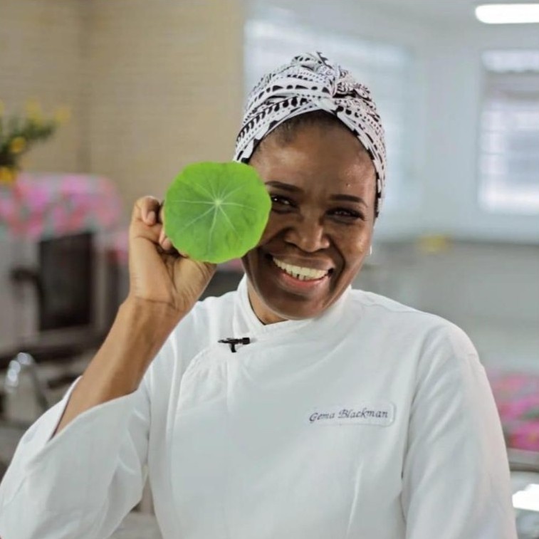 Mulher negra profissional de gastronomia apresentando ingrediente natural em cozinha profissional, destacando culin&aacute;ria artesanal, sustentabilidade e preparo de alimentos frescos.