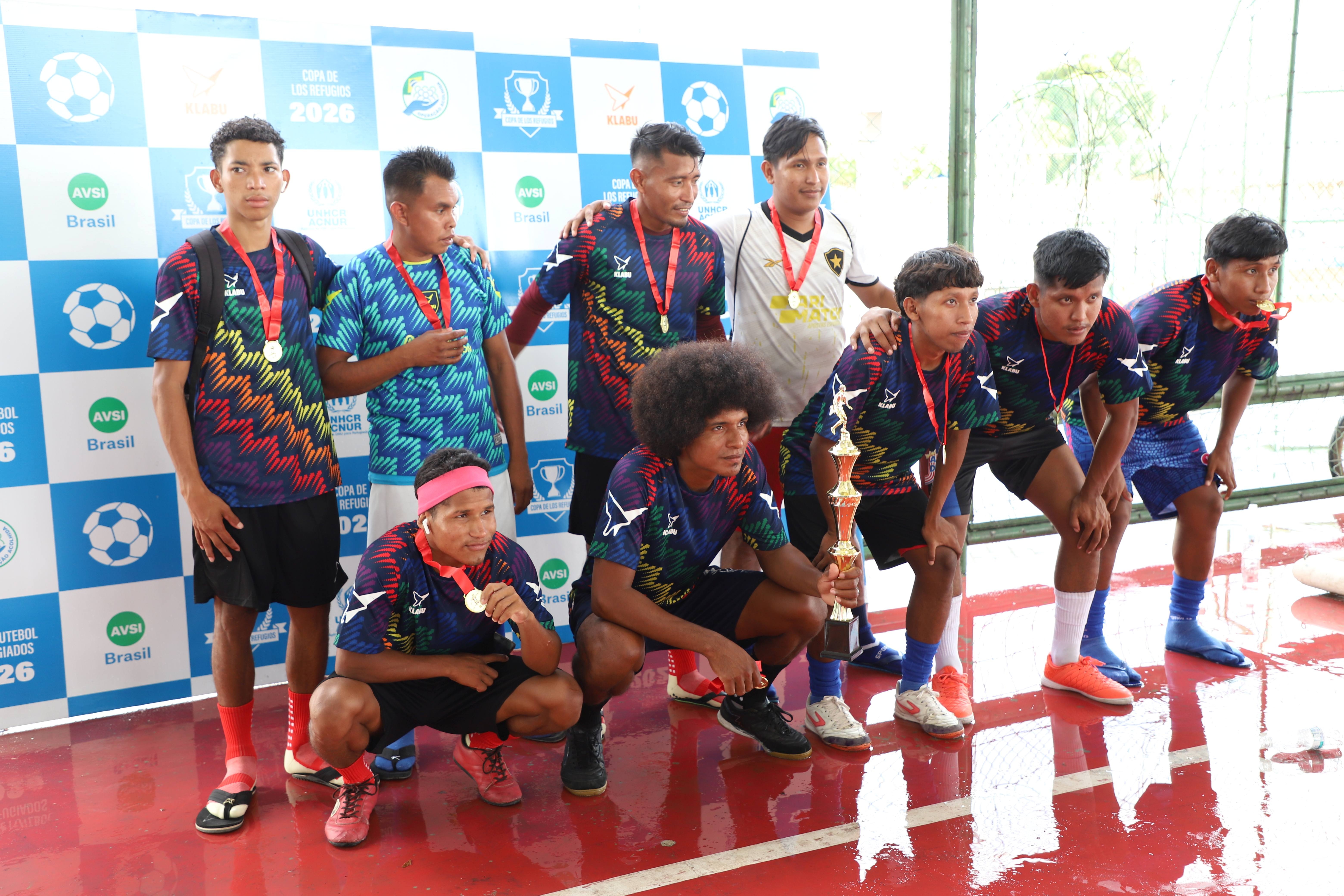 Grupo de nove atletas posa para uma foto em frente a um painel da Copa de los Refugiados 2026, com logotipos da AVSI Brasil, ACNUR e parceiros do evento. Os participantes vestem uniformes esportivos azul-marinho com estampas coloridas e usam medalhas no pesco&ccedil;o. Uma pessoa agachada ao centro segura um trof&eacute;u dourado, enquanto as demais se posicionam ao redor para a foto. A imagem foi registrada em uma quadra esportiva coberta, com piso vermelho e cerca de prote&ccedil;&atilde;o ao fundo.
