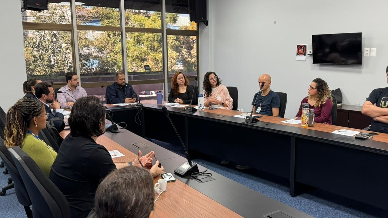 Protocolos de Atendimento Integrado Grupo de aproximadamente dez pessoas reunido em uma sala de reuniões corporativa, sentado ao redor de uma mesa em formato de U. Uma pessoa fala enquanto as demais acompanham a discussão.