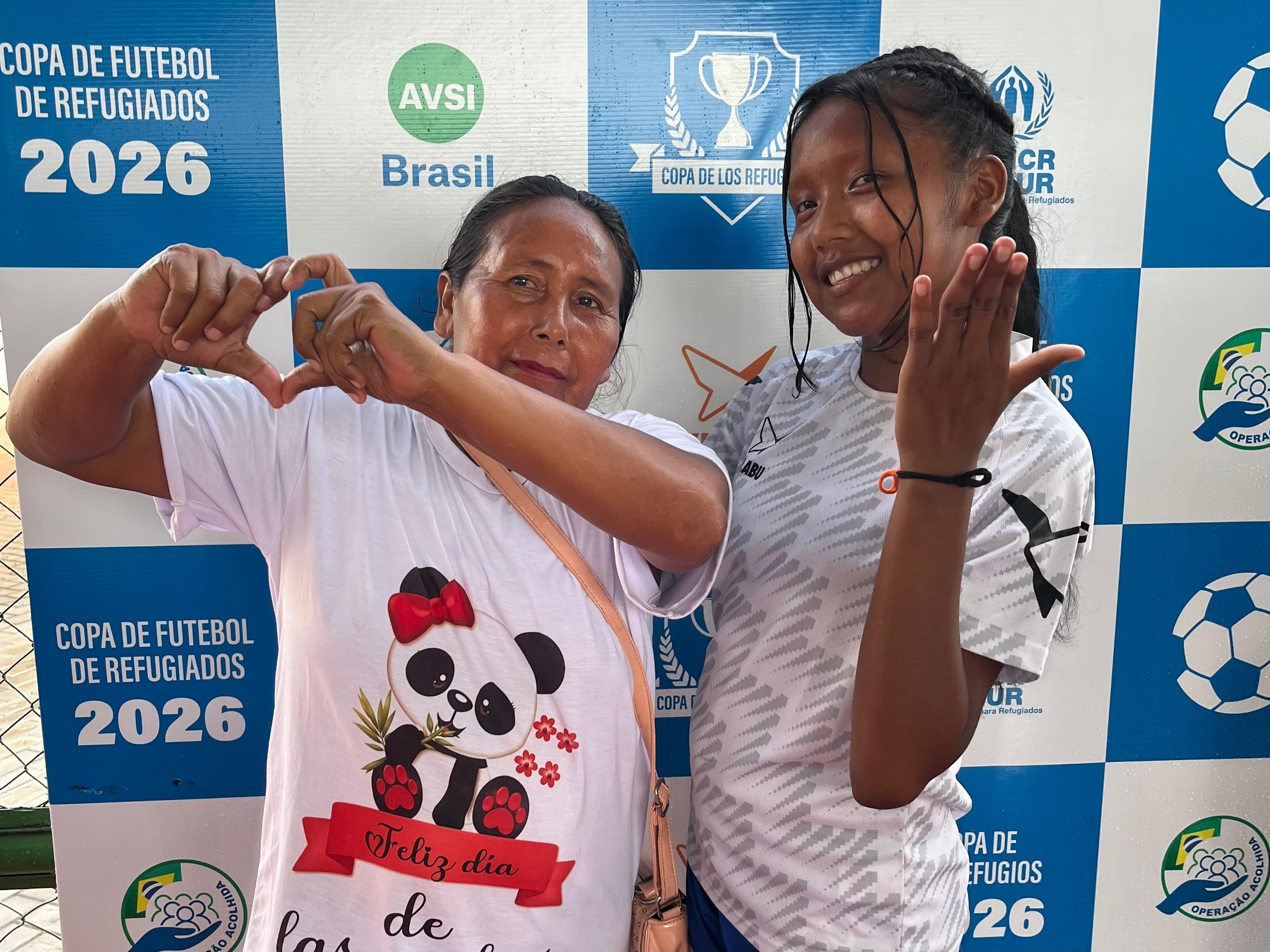 Duas pessoas posam diante de um painel de patrocinadores da Copa de Futebol de Refugiados 2026. Uma pessoa &agrave; esquerda veste uma camiseta branca com a ilustra&ccedil;&atilde;o de um panda e faz um gesto de cora&ccedil;&atilde;o com as m&atilde;os. A pessoa &agrave; direita veste uniforme esportivo branco e azul e faz um gesto com a m&atilde;o em dire&ccedil;&atilde;o &agrave; c&acirc;mera. Ao fundo, h&aacute; um painel quadriculado azul e branco com os logotipos da AVSI Brasil, ACNUR e da Copa de los Refugiados, al&eacute;m de imagens de bolas de futebol.