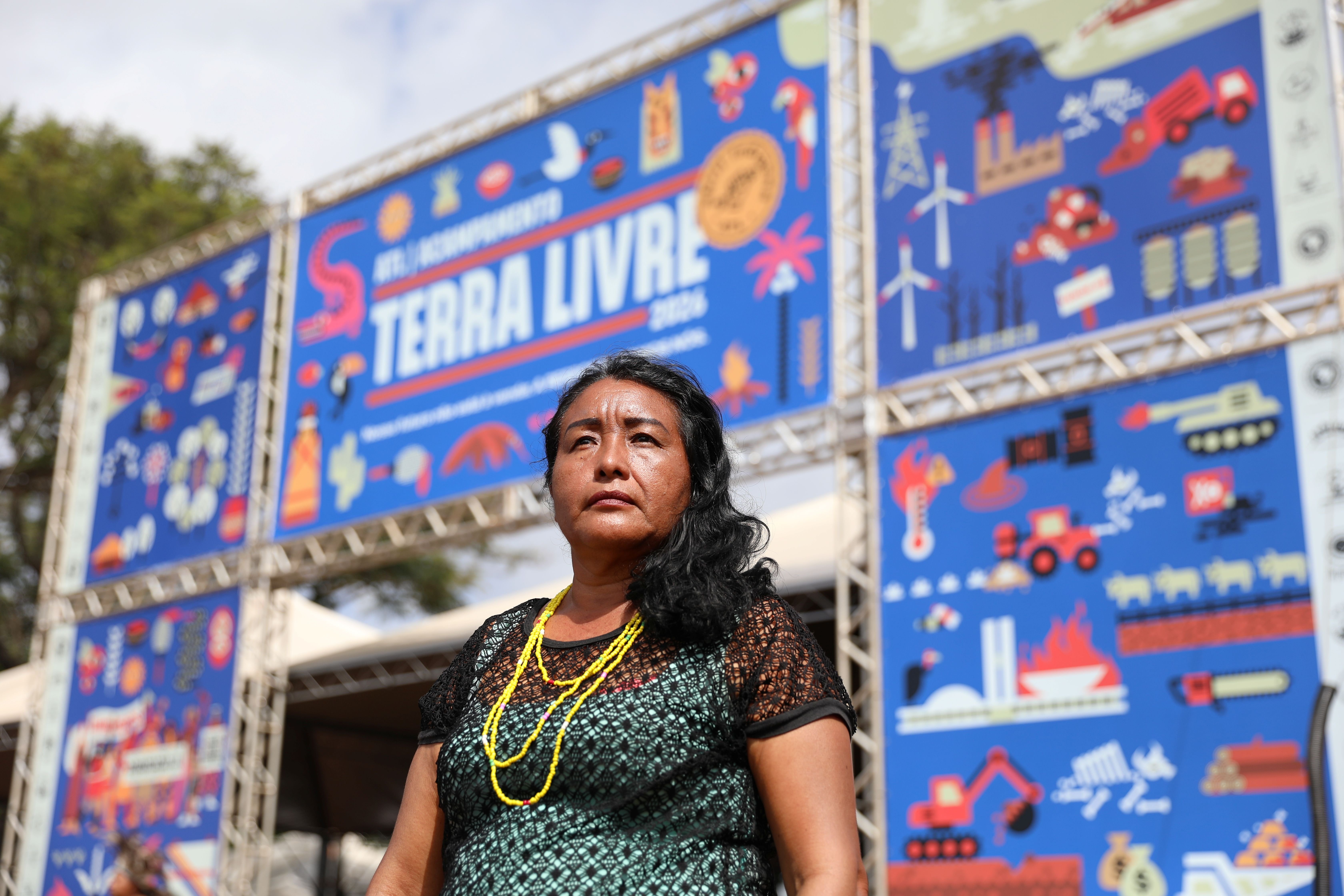 Mulher ind&iacute;gena em primeiro plano diante de um grande painel azul de evento ao ar livre. O painel exibe o texto &ldquo;ATL Acampamento Terra Livre&rdquo; e diversas ilustra&ccedil;&otilde;es relacionadas a meio ambiente, energia, ind&uacute;stria e territ&oacute;rio. A pessoa usa colares de contas amarelas e est&aacute; posicionada no centro da imagem, com a estrutura do evento e &aacute;rvores ao fundo.