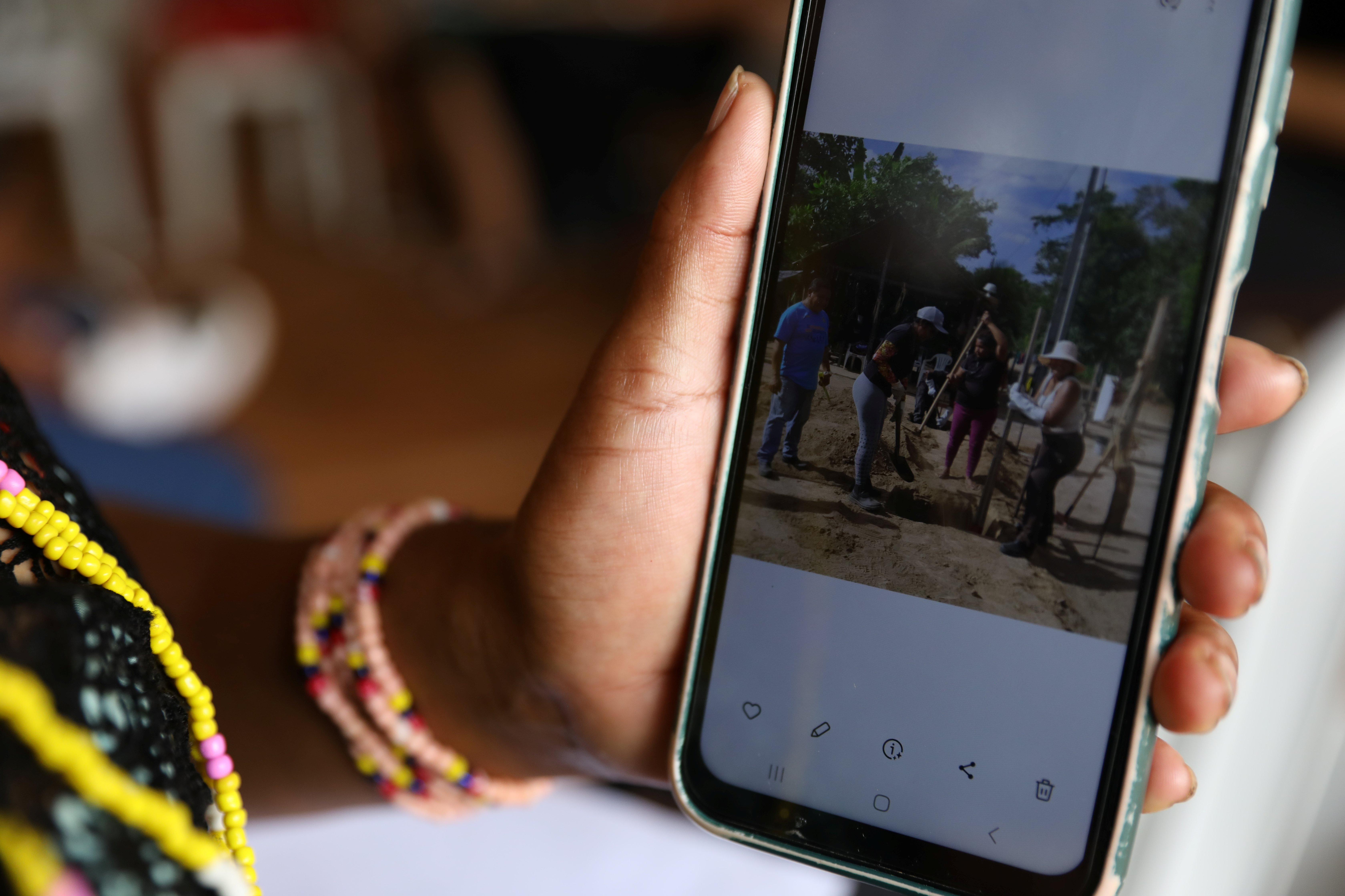 Pessoa segura um smartphone exibindo uma foto de um grupo reunido ao ar livre em uma &aacute;rea de solo arenoso. Em primeiro plano, s&atilde;o vis&iacute;veis a m&atilde;o que segura o aparelho e pulseiras coloridas no pulso. O fundo da cena est&aacute; desfocado, destacando a tela do celular.