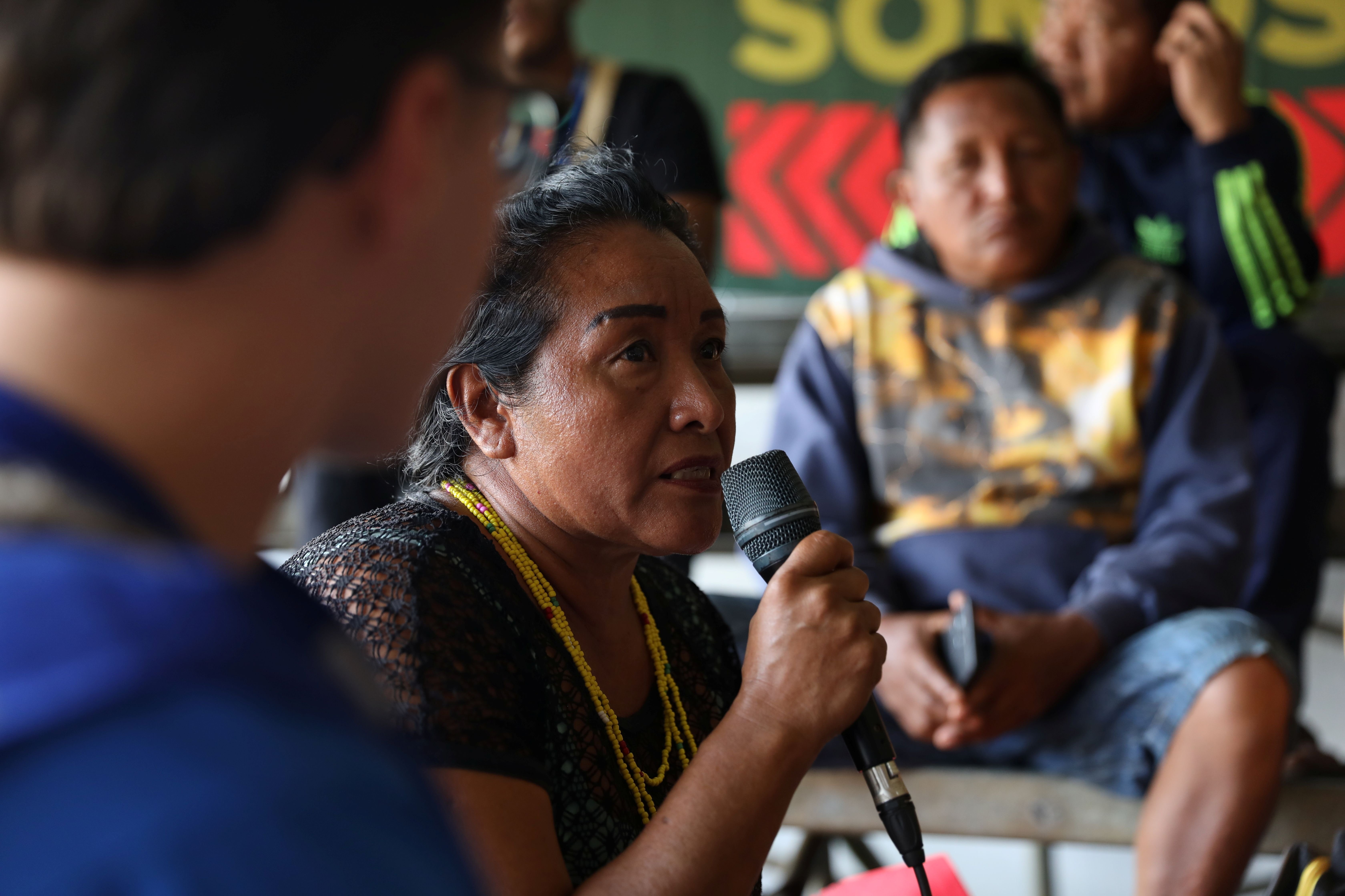 Alida participa de uma reuni&atilde;o segurando um microfone e falando enquanto est&aacute; sentada em um ambiente coletivo. A imagem destaca Alida, quem est&aacute; com a palavra, usando colares de contas amarelas, enquanto outras pessoas aparecem desfocadas ao redor e ao fundo, acompanhando a discuss&atilde;o.