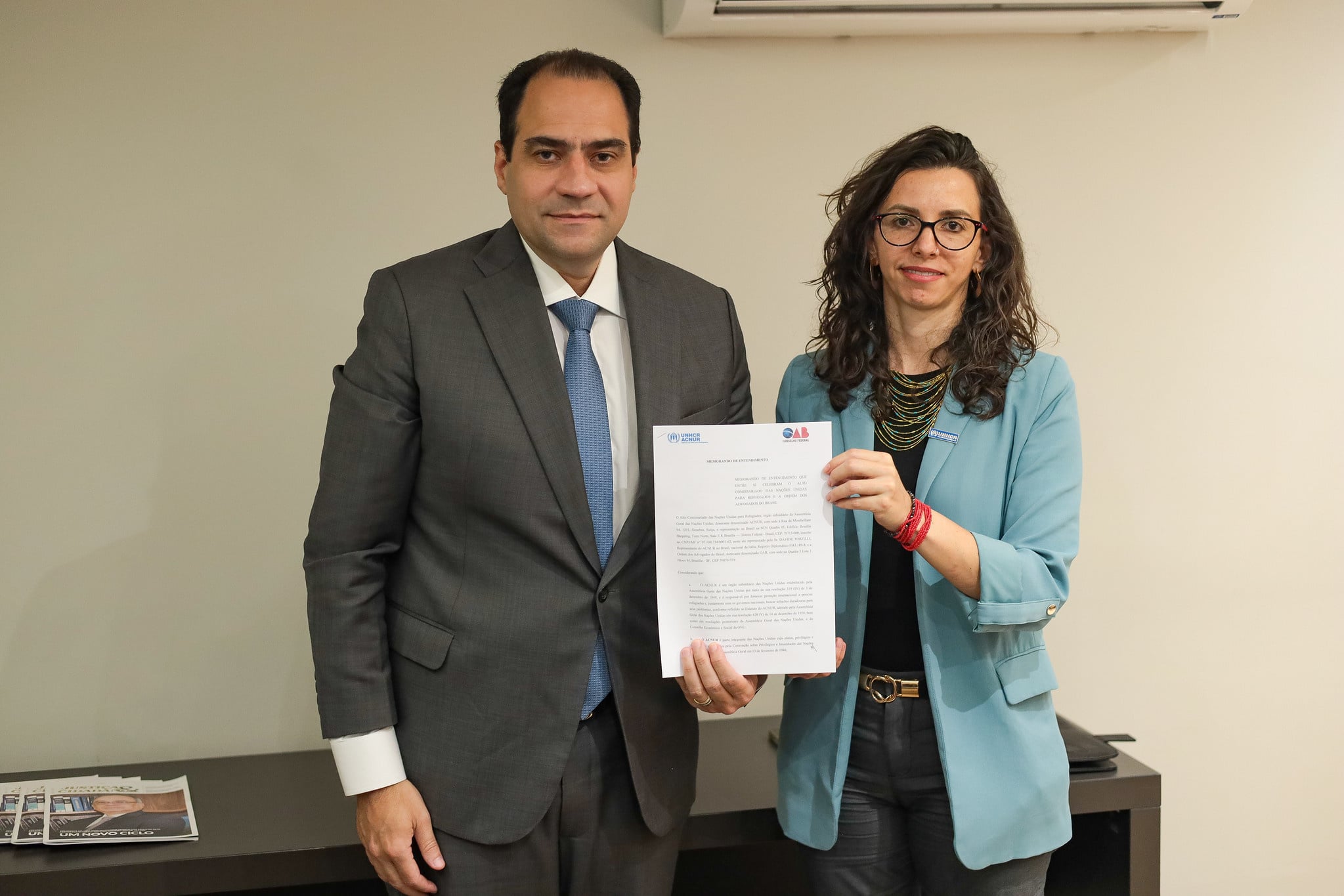 Representantes do ACNUR e da OAB exibem documento assinado durante formaliza&ccedil;&atilde;o de parceria institucional em escrit&oacute;rio corporativo.