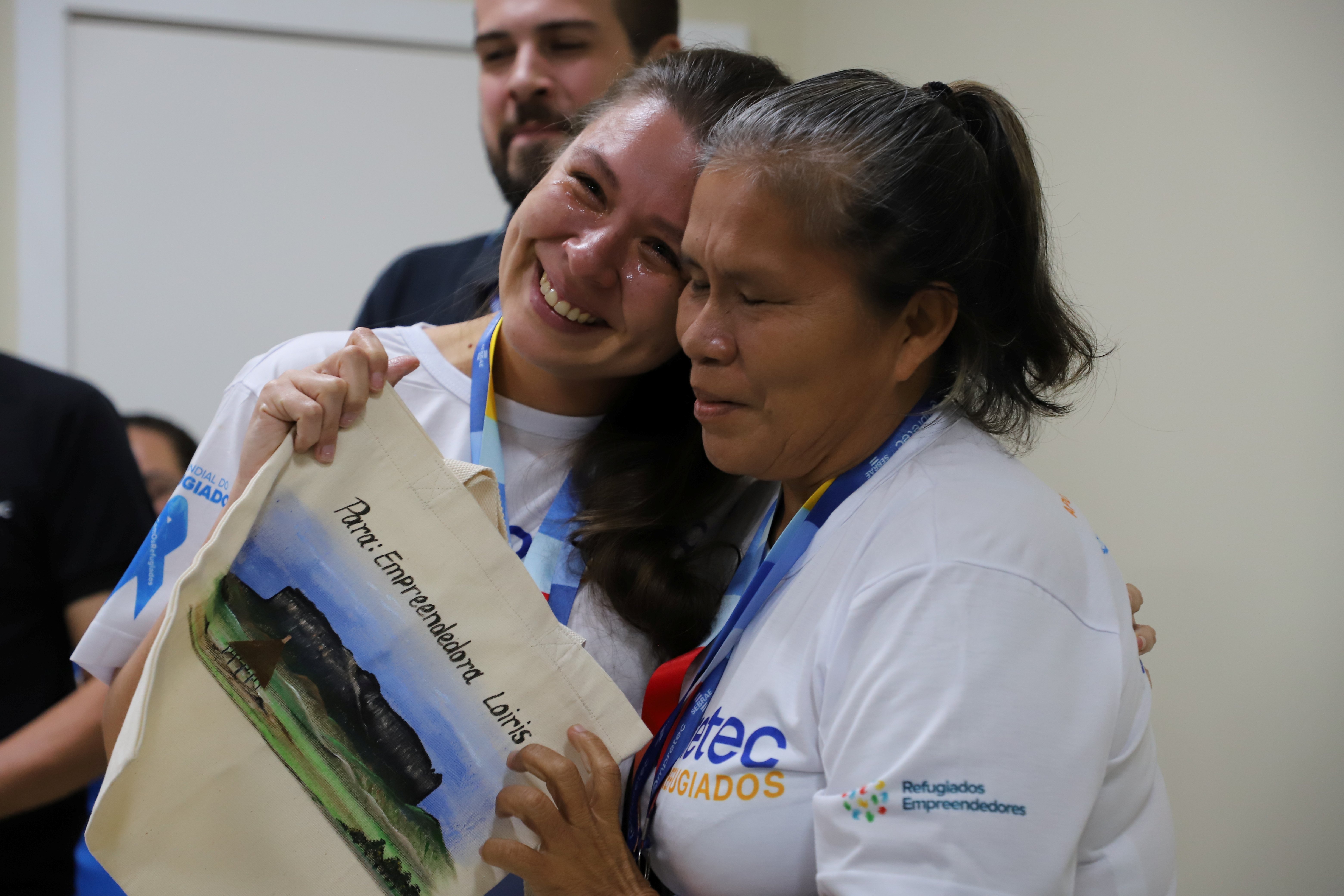 Duas participantes do programa Empretec para Refugiados compartilham um abra&ccedil;o durante atividade de encerramento, enquanto uma delas segura uma bolsa personalizada que ganhou de presente.