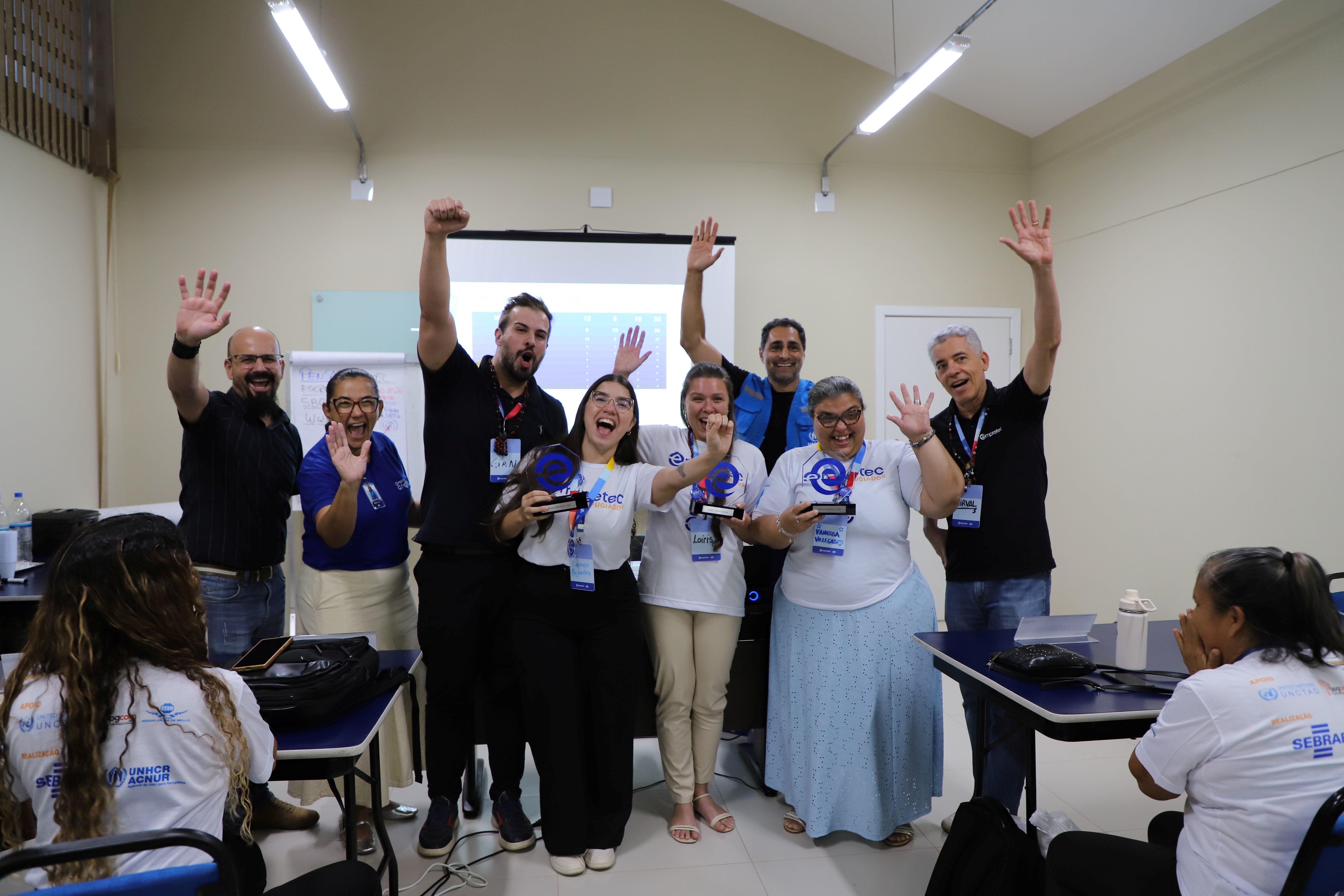 Participantes, facilitadores e representantes de organiza&ccedil;&otilde;es parceiras celebram a conclus&atilde;o do programa Empretec para Refugiados em sala de treinamento, com trof&eacute;us e certificados apresentados aos concluintes.