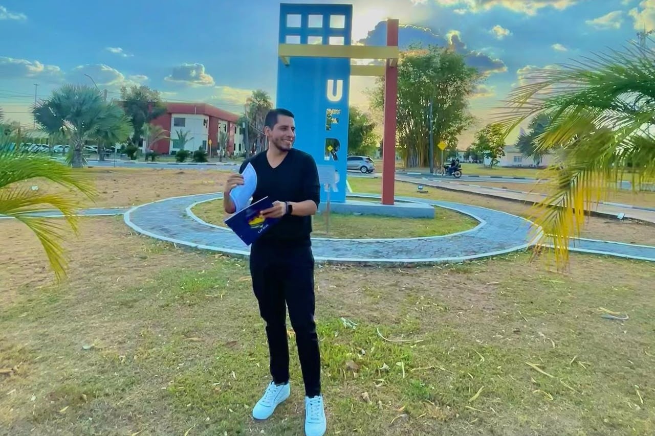 Franco segura seu diploma revalidado pela Universidade Federal de Roraima em um gramado com um monumento de concreto atr&aacute;s com as iniciais da UFRR
