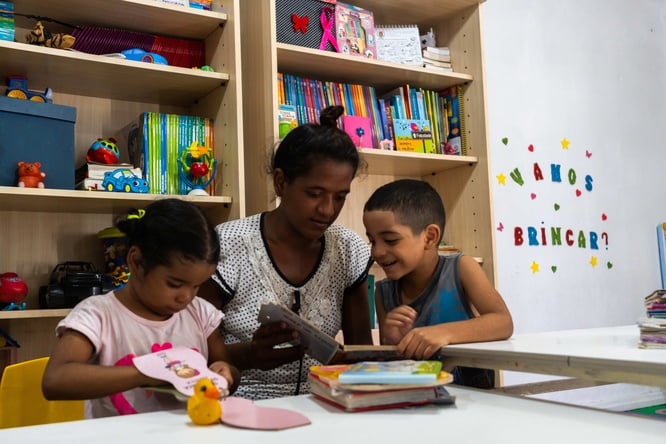 Crianças e uma pessoa adulta folheiam livros em um espaço de leitura com estantes repletas de materiais educativos e brinquedos.