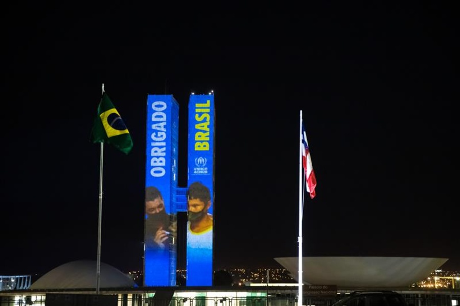 As duas torres do Congresso Nacional do Brasil estão iluminadas à noite e exibem as palavras 'OBRIGADO BRASIL' em letras grandes, com 'OBRIGADO' em azul no prédio da esquerda e 'BRASIL' em amarelo no da direita. Imagens de pessoas são projetadas nas fachadas. Em frente, há dois mastros com bandeiras — a do Brasil à esquerda e outra não identificada à direita. O céu está escuro e há luzes da cidade ao fundo."