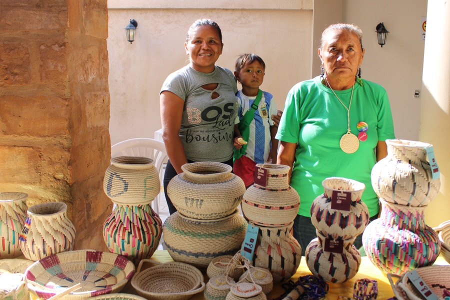 Três pessoas indígenas em pé, sendo duas mulheres e uma criança ao meio, atrás de uma mesa com cestos e itens artesanais de diferentes formas, tamanhos e cores, exibindo padrões detalhados. 