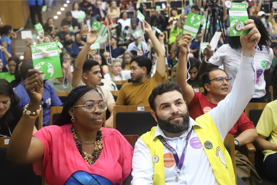 Auditório cheio de pessoas participando da COMIGRAR, todas sentadas em fileiras e levantando cartões de votação. No primeiro plano, duas pessoas seguram os cartões, uma delas usando um colete amarelo com crachá e adesivos e outra ao lado de blusa rosa também levantando um cartão.