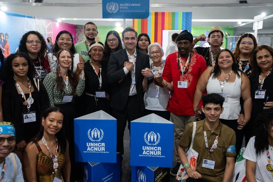 Grupo de pessoas reunidas em um estande da ONU com dois totens azuis exibindo o logotipo do ACNUR (Agência da ONU para Refugiados). Ao fundo, há um painel com faixas coloridas e outro com o emblema das Nações Unidas. Algumas pessoas usam crachás e seguram sacolas, indicando participação na COP30.