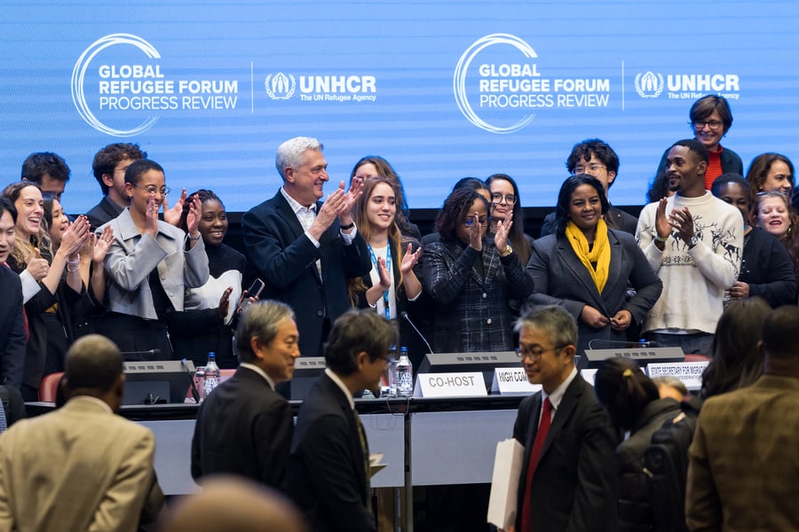 Grupo diverso de participantes aplaude no palco durante a sessão de encerramento da Revisão de Progresso do Fórum Global sobre Refugiados. Ao fundo, um painel azul exibe os logotipos do ACNUR e do Fórum Global sobre Refugiados. Em primeiro plano, representantes e delegados circulam pelo espaço da conferência, com mesas, microfones e placas institucionais visíveis, reforçando o caráter formal e internacional do evento.
