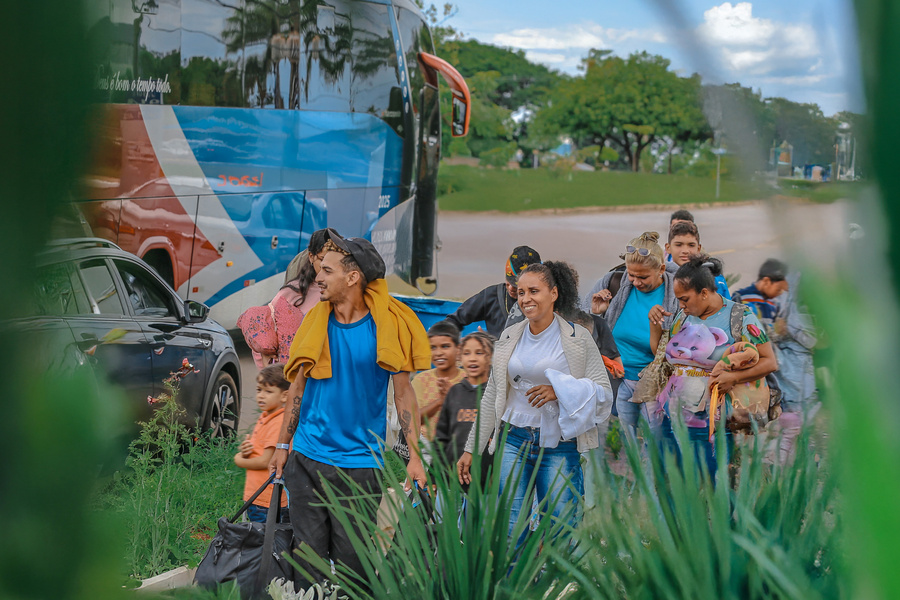 Grupo de pessoas caminhando com malas e crianças próximo a um ônibus estacionado, em uma área aberta com vegetação em primeiro plano e árvores ao fundo.