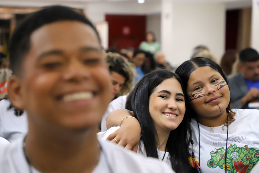 Crianças sentadas em um auditório. Em primeiro plano, uma criança aparece desfocada. Ao fundo, duas crianças próximas uma da outra olham em direção à câmera, vestindo camisetas claras com ilustrações. Há outras pessoas sentadas atrás, em ambiente interno bem iluminado.