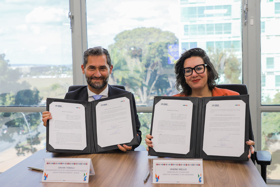 Representante do ACNUR e Ministra dos Direitos Humanos no Brasil estão sentados segurando um acordo assinado nas mãos e sorrido para a foto