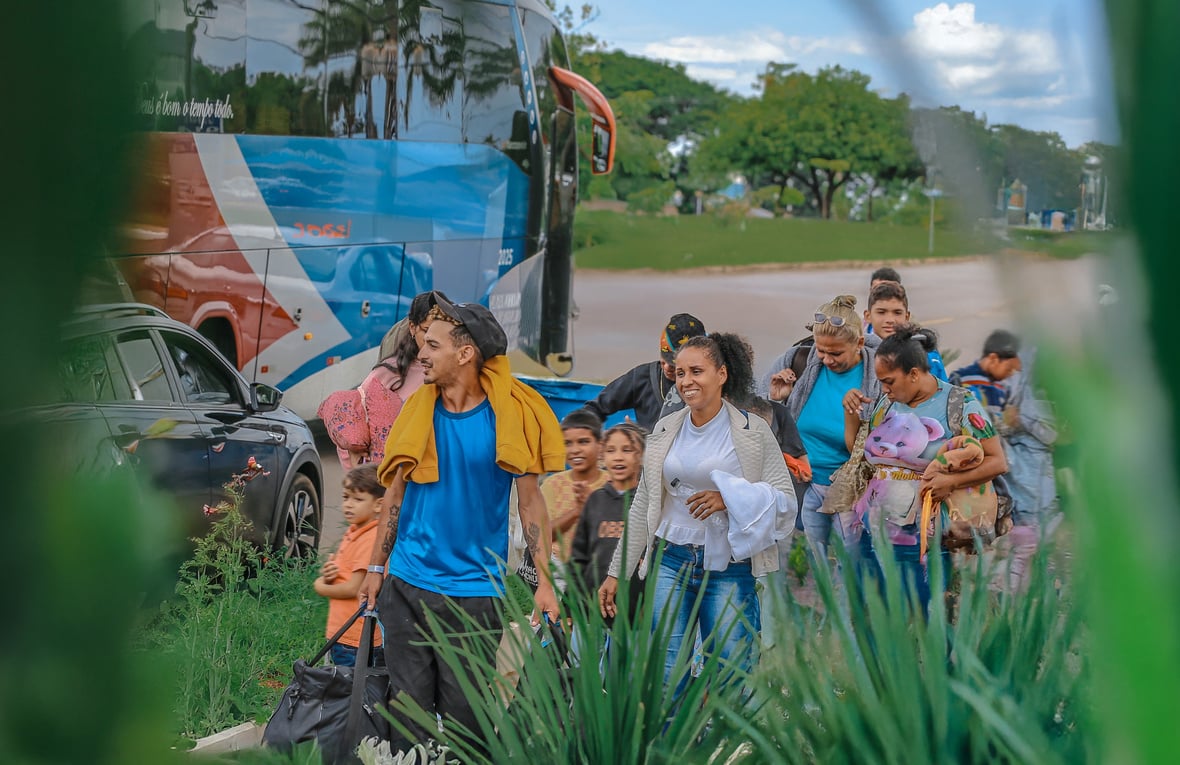 Grupo de pessoas caminhando com malas e crianças próximo a um ônibus estacionado, em uma área aberta com vegetação em primeiro plano e árvores ao fundo.
