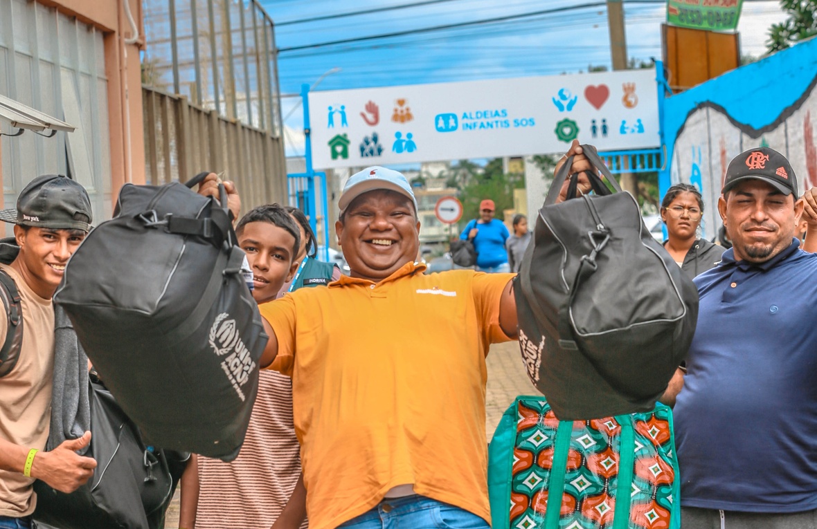 Grupo de pessoas carregando mochilas e bolsas grandes com a logomarca do ACNUR, enquanto caminham por uma área externa. Ao fundo, há um portão com a sinalização da organização ‘Aldeias Infantis SOS’. As pessoas parecem estar em movimento, algumas levantando as bagagens, com construções e um mural colorido ao redor.