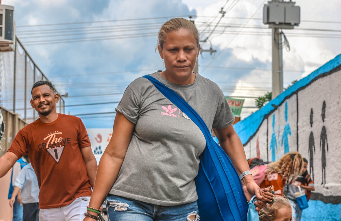 Uma mulher com a expressão facial fechada caminha em uma rua movimentada, carregando uma sacola na mão e uma bolsa azul transversal. Ela segura a mão de uma criança. Em segundo plano, um homem também caminha sorrindo. O ambiente é urbano, com muros pintados, fiação elétrica aérea e um céu parcialmente nublado. No fundo, há um mural com figuras humanas estilizadas pintadas na parede.