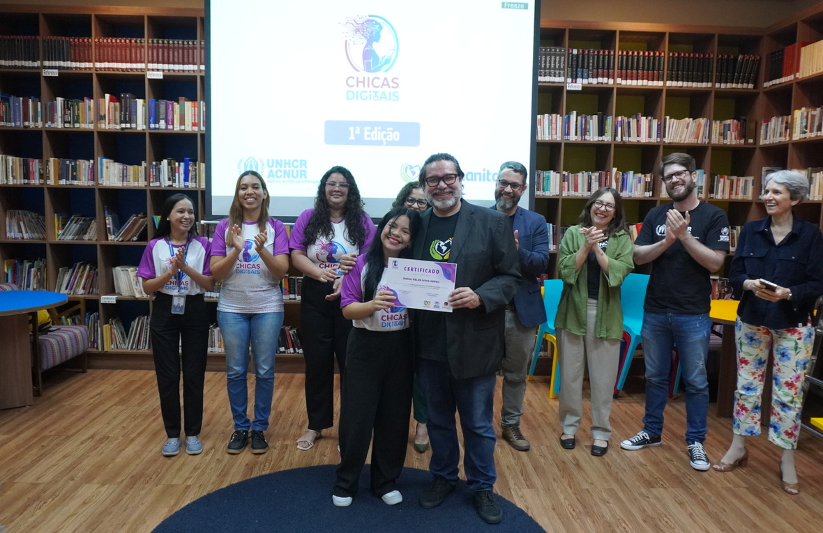 Grupo de pessoas reunidas em uma biblioteca durante um evento do programa ‘Chicas Digitais’. No centro, duas pessoas seguram um certificado. Outras pessoas ao fundo aplaudem. Atrás do grupo há uma tela de projeção exibindo o logotipo do evento e estantes cheias de livros.