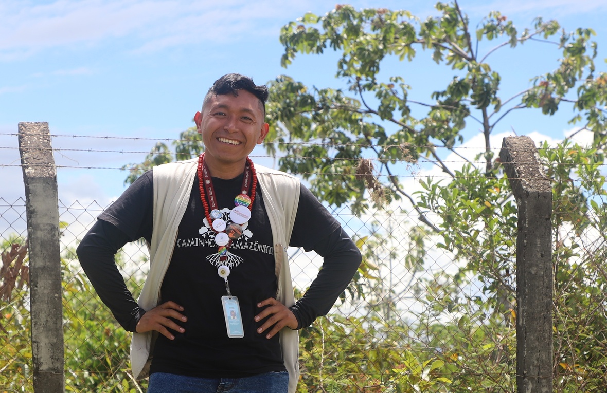 Pessoa em ambiente rural com vegetação e cerca de arame, usando colete claro, camiseta preta e crachá, em registro institucional ao ar livre.