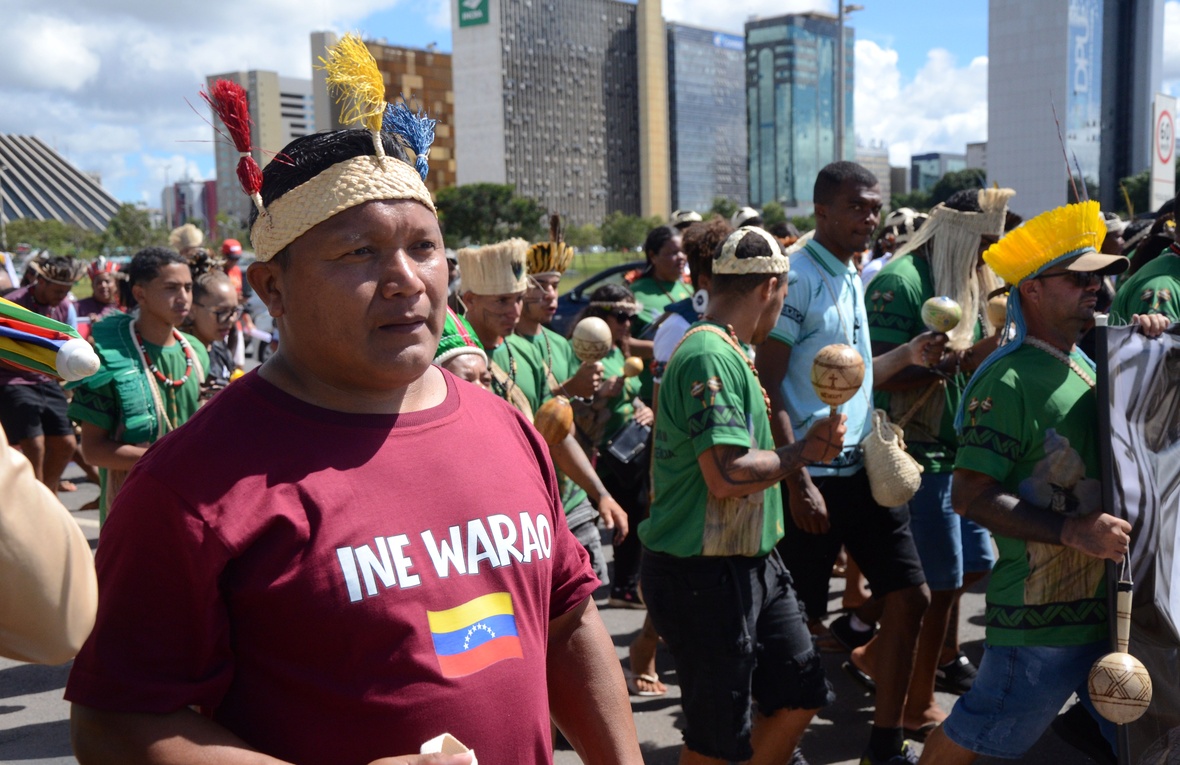 Homem indígena em primeiro plano usa camiseta vinho com o texto ‘INE WARAO’ e uma pequena bandeira estampada, caminhando em meio a um grupo em marcha. Ao redor, várias pessoas vestem camisetas verdes, seguram instrumentos e adornos, com prédios altos ao fundo em uma área urbana.