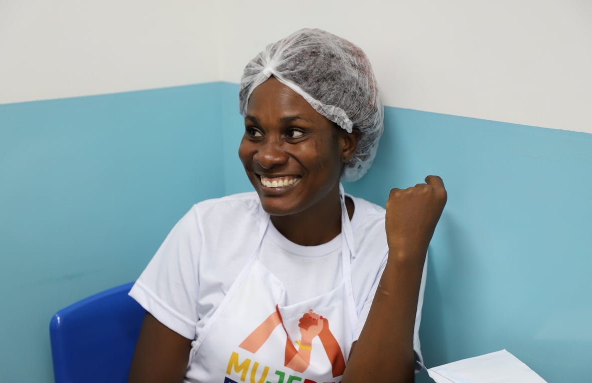 Mulher está sentada frente a uma parede azul e branca. Posa para a foto sorrindo. Ela está vestida com uma camiseta branca com a marca do projeto Mujeres Fuertes. Ela leva touca no cabelo. 