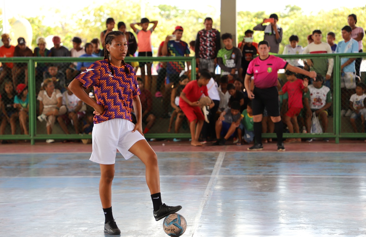 Pessoa participante de uma partida de futsal em uma quadra coberta, com um pé apoiado sobre a bola próximo à linha central. A pessoa veste camisa estampada em laranja e roxo, shorts brancos, meias pretas e chuteiras pretas. Ao fundo, atrás de uma grade verde, há um grande público acompanhando o jogo. Na lateral da quadra, outra pessoa com uniforme rosa está posicionada próxima à área de jogo. A quadra é iluminada pela luz natural, com árvores visíveis ao fundo.