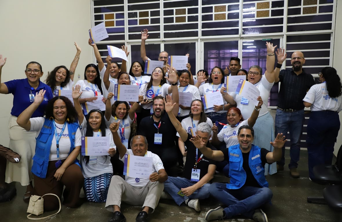 Grupo de participantes do programa Empretec para Refugiados posa com certificados ao final da capacitação, acompanhado por representantes de organizações parceiras, em sala de treinamento.