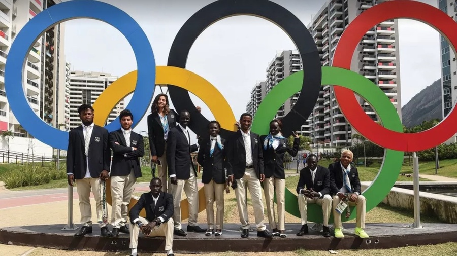 Grupo de pessoas vestidas com trajes formais posando em frente à escultura dos anéis olímpicos, com prédios altos e céu claro ao fundo.