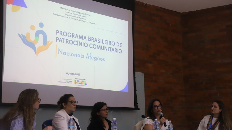Painel com cinco pessoas sentadas à mesa com microfones e garrafas de água, diante de uma tela que exibe slide sobre o Programa Brasileiro de Patrocínio Comunitário para Nacionais Afegãos, apresentado pelo Ministério da Justiça e Segurança Pública.