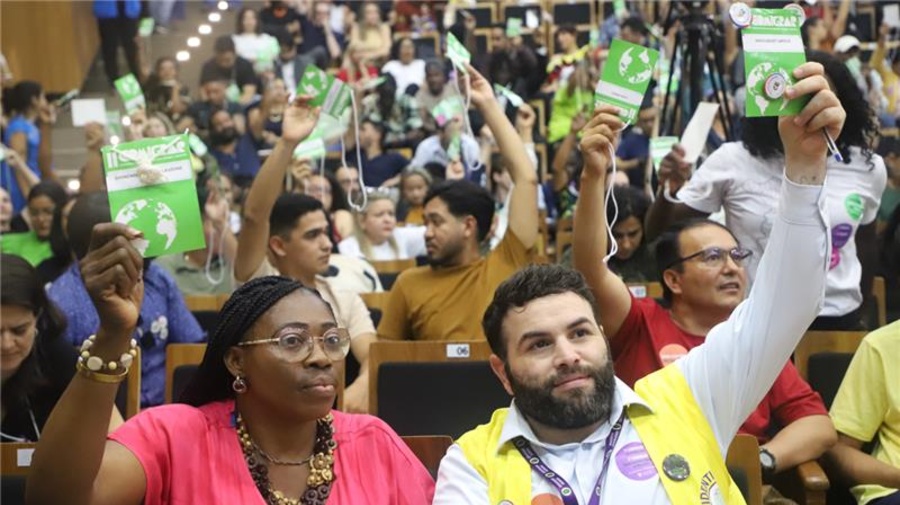 Auditório cheio de pessoas participando da COMIGRAR, todas sentadas em fileiras e levantando cartões de votação. No primeiro plano, duas pessoas seguram os cartões, uma delas usando um colete amarelo com crachá e adesivos e outra ao lado de blusa rosa também levantando um cartão.