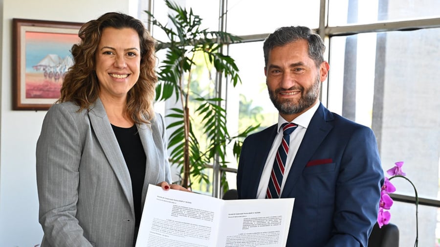 Duas pessoas em ambiente institucional seguram documento oficial aberto após assinatura, com plantas e janelas ao fundo