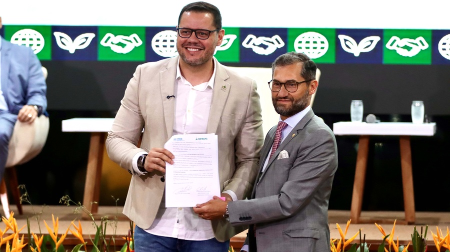 Entrega oficial de documento durante evento institucional, com participantes no palco e painel de identidade visual ao fundo.