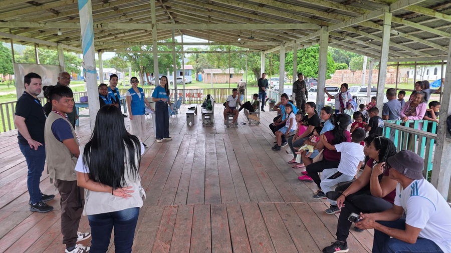 Grupo de pessoas sentadas e em pé em um espaço comunitário coberto, participando de uma atividade coletiva mediada por equipes de apoio, com casas e árvores ao fundo.
